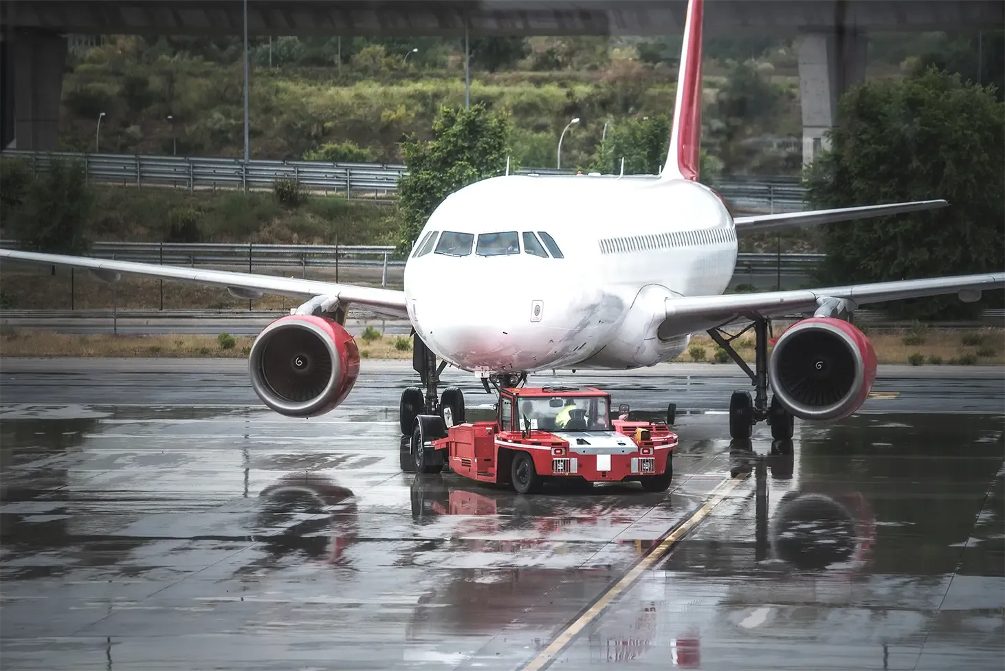 Red pushback vehicle towing a commercial airplane on a wet runway.
