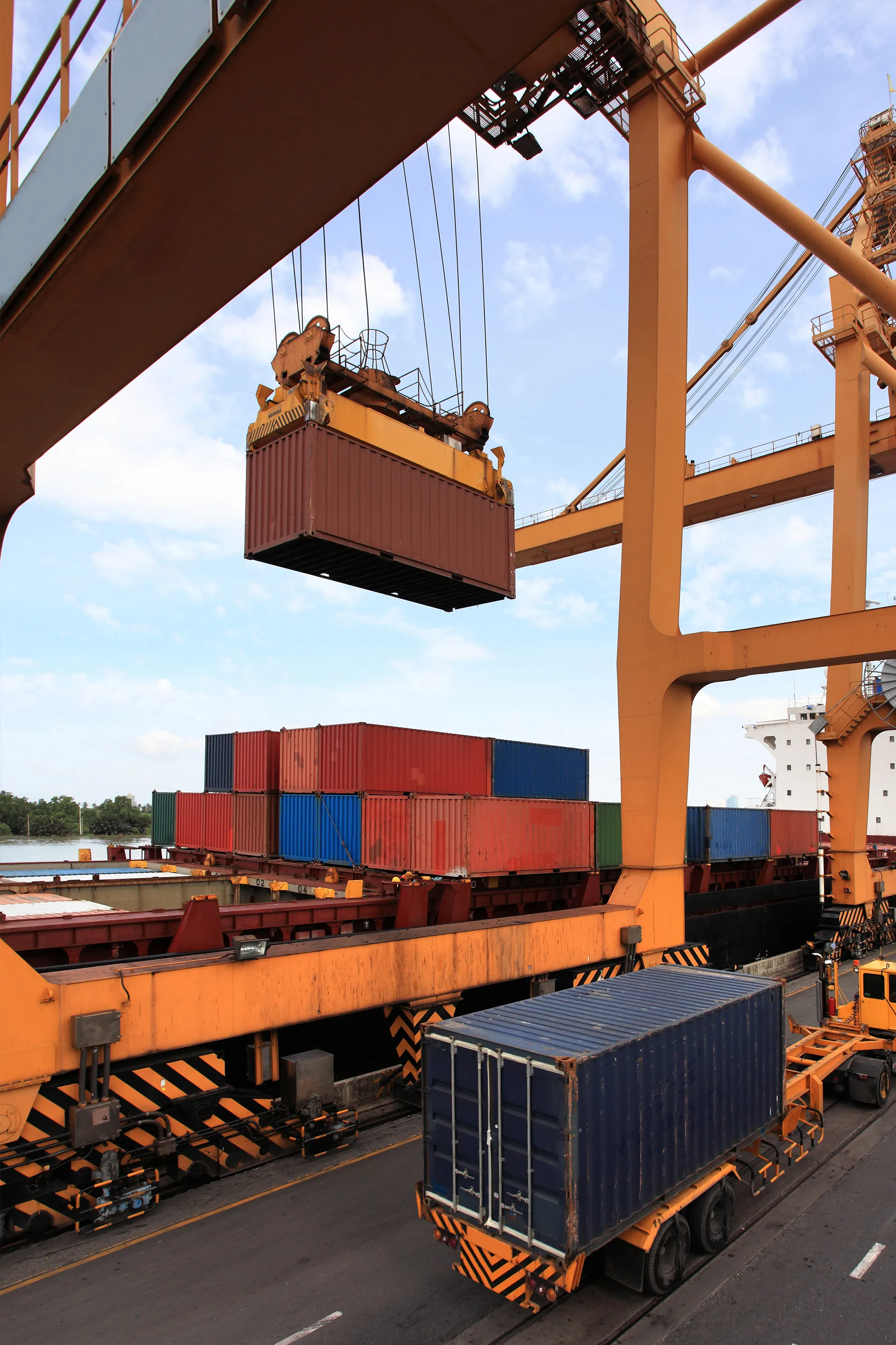 Grua pórtico naranja moviendo un contenedor marrón hacia un buque de carga.