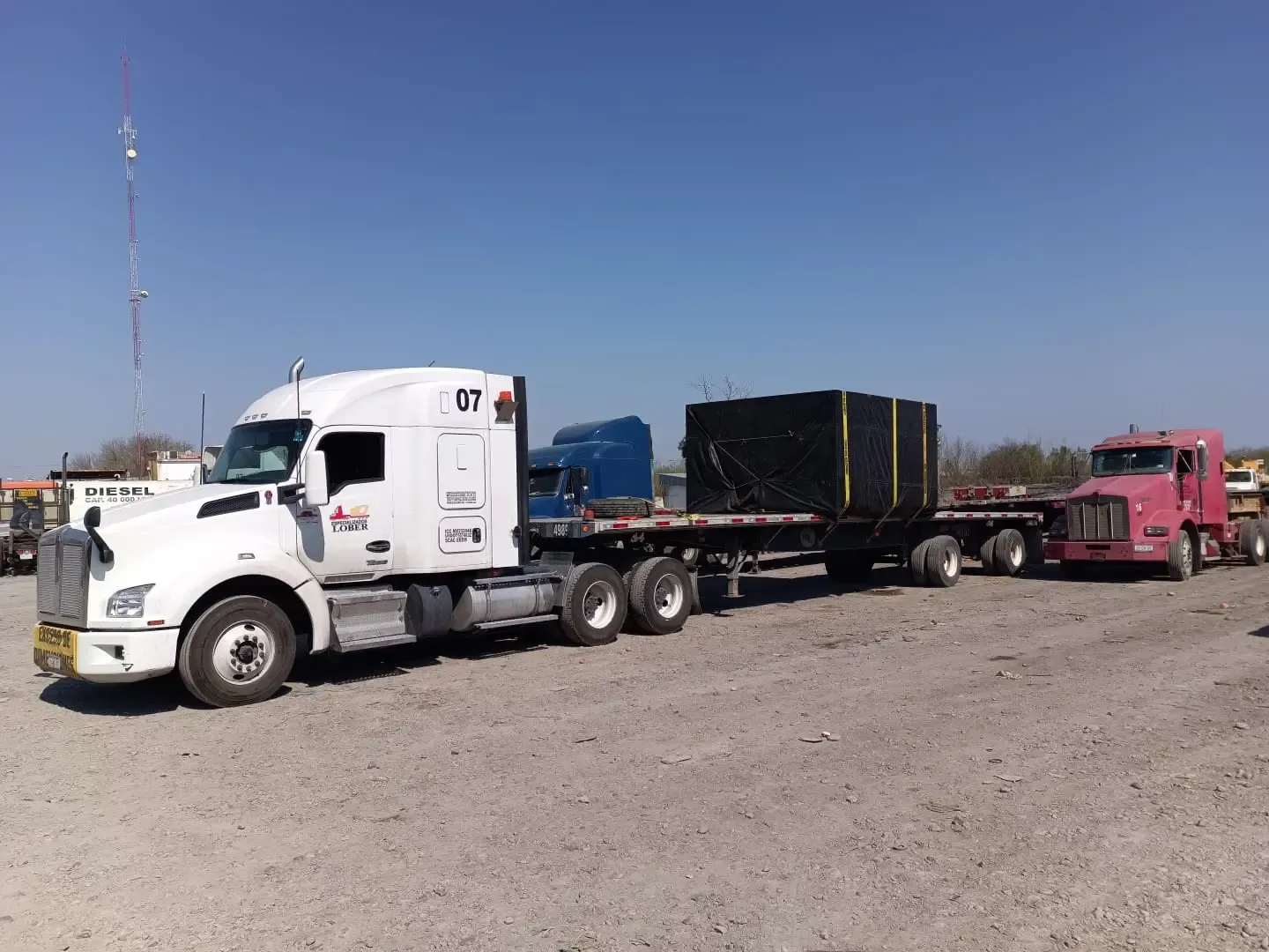 Convoy de camiones de carga pesada transportando mercancía con exceso de dimensión en terreno abierto.