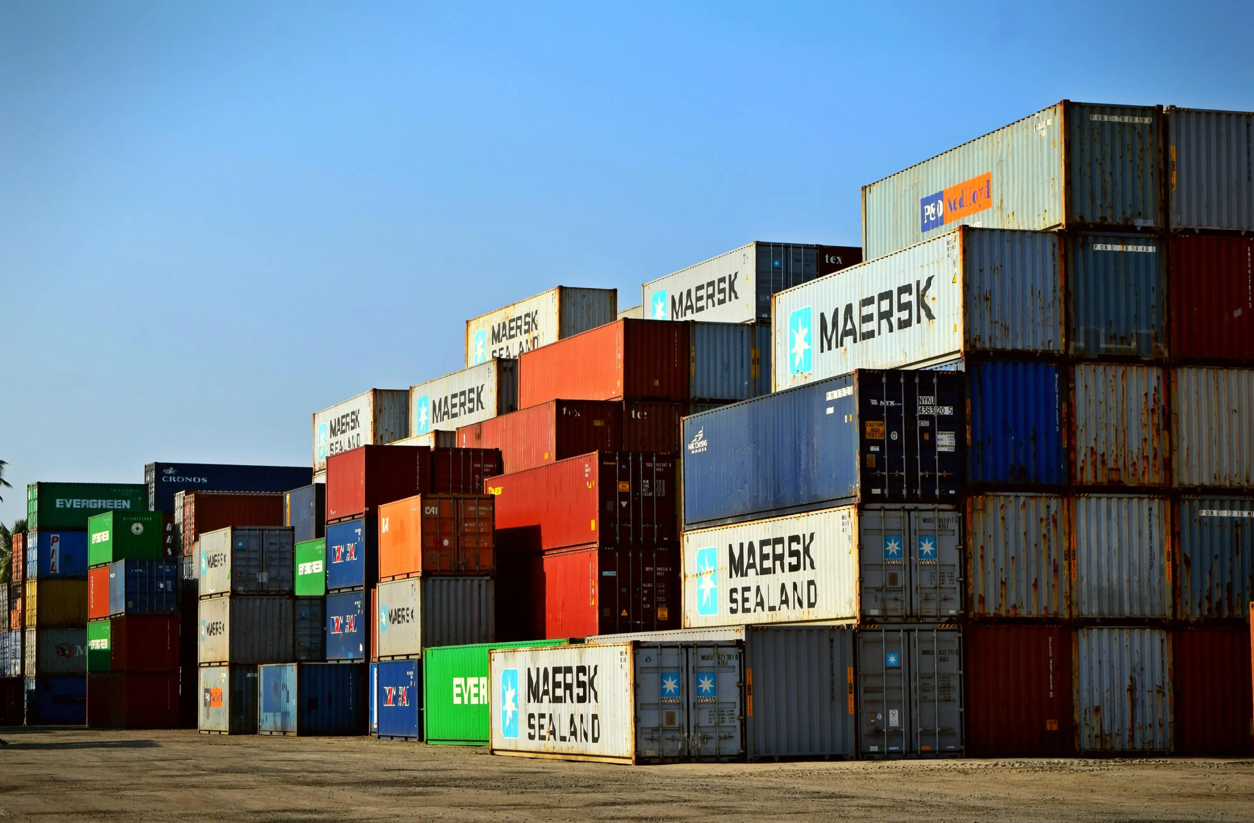 Port terminal infrastructure and intermodal transport Long row of stacked containers at a river pier with cargo cranes under a cloudy sky.