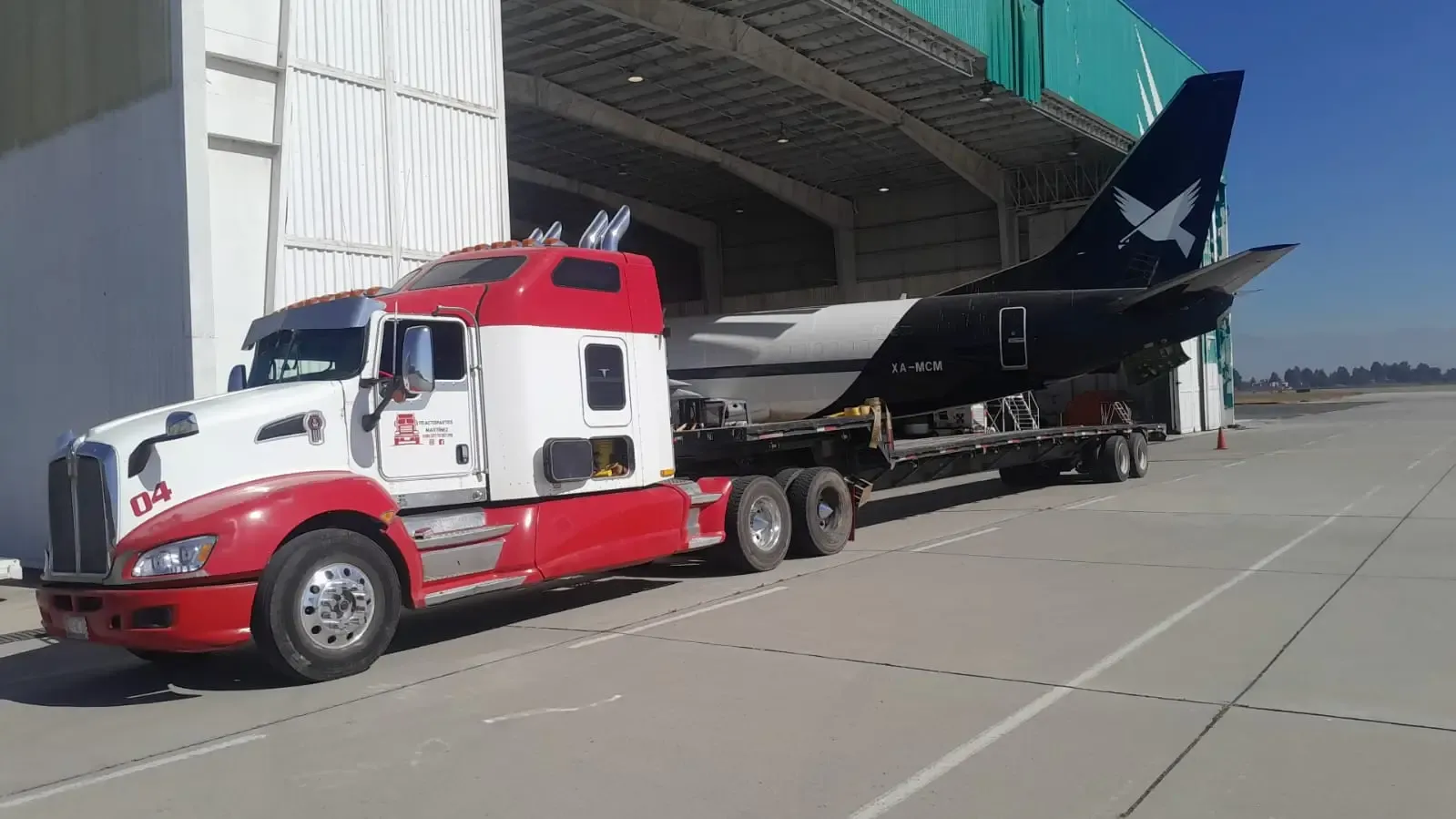 Red and white Kenworth truck transporting an aircraft fuselage tail number XA-MCM on a lowboy trailer outside a hangar.